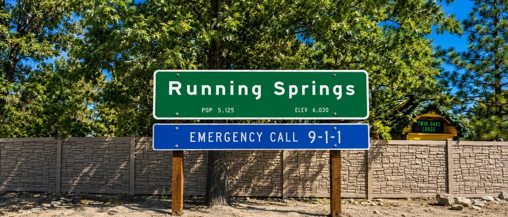 Wide view of the Running Springs community sign on wooden posts, reading Running Springs, pop 5,125, elev 6,030, with an emergency call 9-1-1 plaque below, trees, a stone wall, and Twin Oaks Lodge visible to the right.
