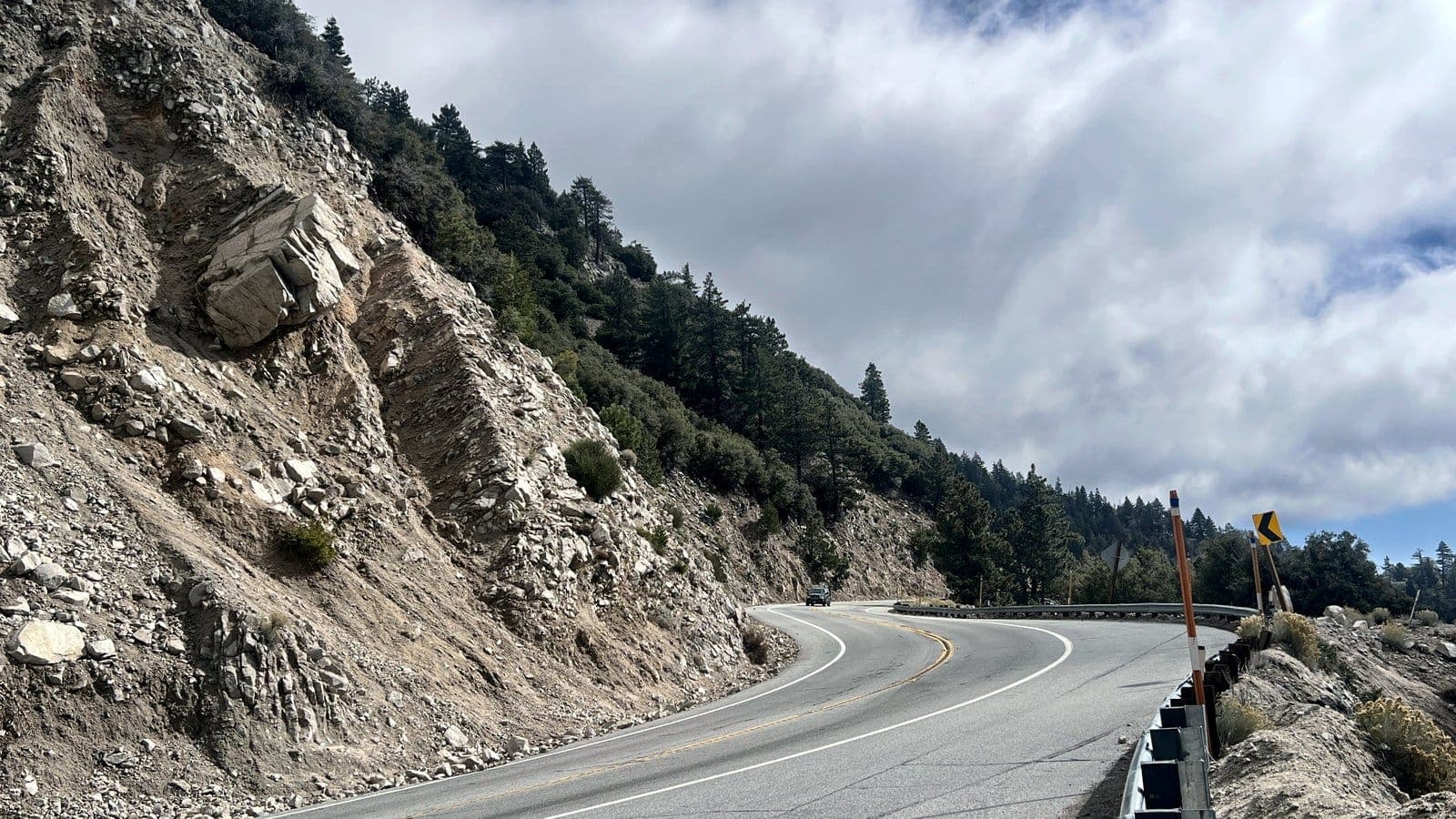 A curving mountain road with guardrail beneath rocky cliffs and cloudy skies in the San Bernardino Mountains