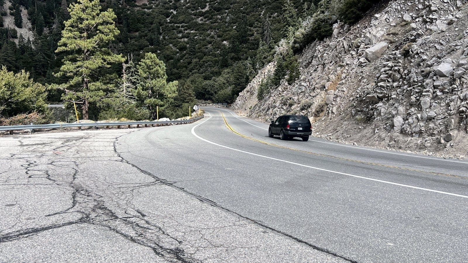 A dark SUV rounds a curve on a mountain highway past a granite rock cut in the San Bernardino Mountains