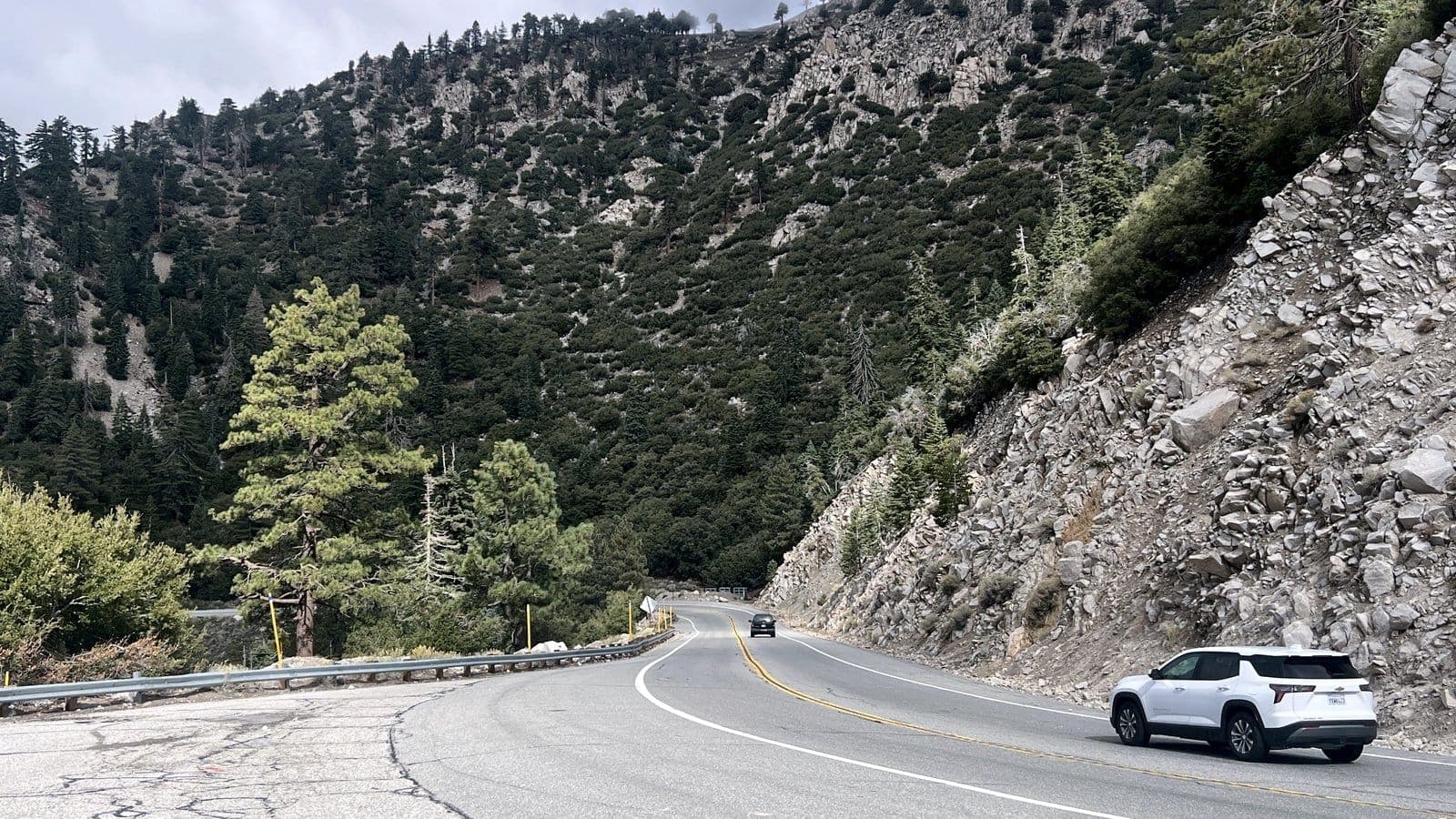 A white SUV drives a winding mountain highway beside granite cliffs and pines in the San Bernardino Mountains