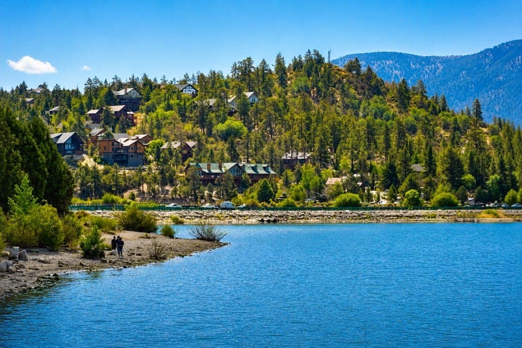 A rocky shoreline, pines, and large homes along a mountain lake under a clear sky.